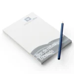 Bloc de notas con Logo de Empresa, mostrando un diseño minimalista con el logotipo de ImprentaPro en la esquina superior derecha. A la izquierda, un lápiz azul inclinado acompaña al bloc, que está parcialmente enrollado en la parte superior