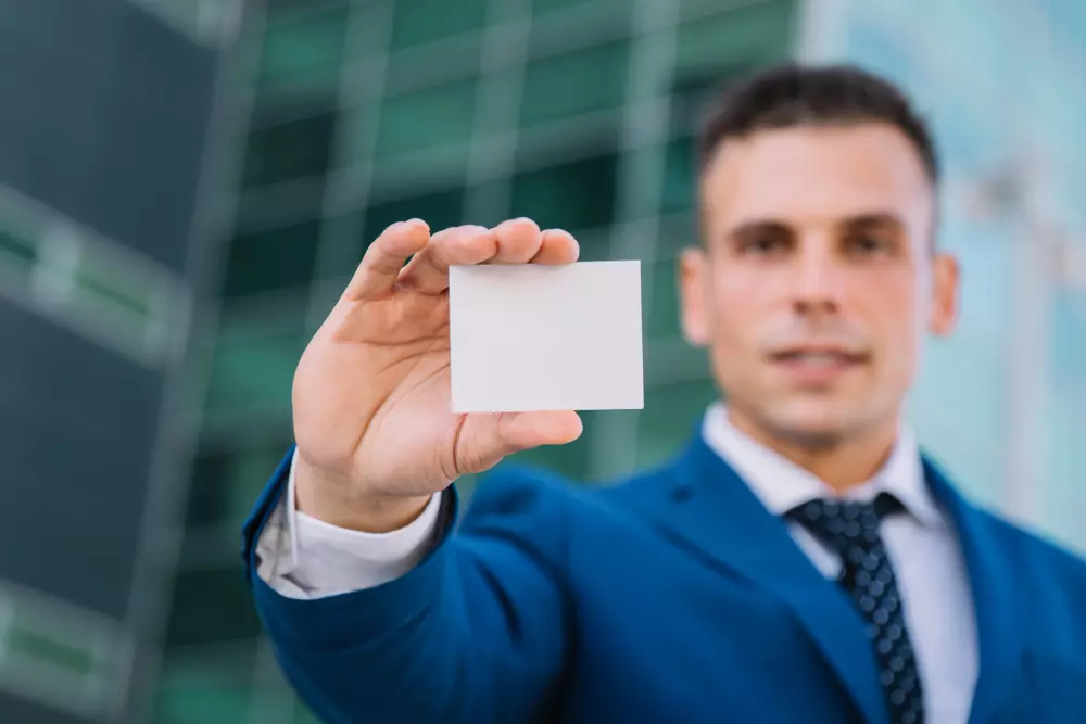 Hombre de negocios con traje azul sosteniendo una tarjeta de visita en blanco hacia la cámara, destacando la importancia del diseño de tarjetas de presentación profesionales en entornos corporativos. La imagen simboliza el valor de las tarjetas de visita como herramienta para causar una buena primera impresión en el mundo empresarial.